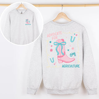 Light gray crewneck sweatshirt with a colorful design featuring a pink cowboy boot tied with a teal bow, surrounded by stars, horseshoes, and a small teal tractor. The text reads “Advocate for Agriculture” in pink and teal lettering.
